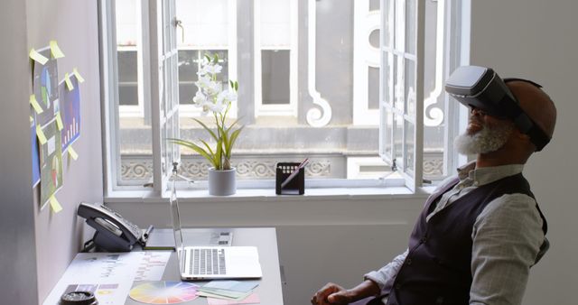 Businessman sitting at office desk using virtual reality headset. He appears relaxed and immersed in the VR experience. Ideal for use in articles or advertisements about modern technology in the workplace, VR applications in business, or promoting innovative work environments.