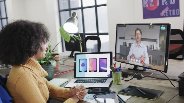 Depicts an African American businesswoman engaging in a video call with a male colleague on screen. The modern office desk includes laptops and design tools, symbolizing a dynamic business environment. Ideal for illustrating remote work scenarios, professional communication, coworking environments, and international teamwork in presentations, websites, or promotional materials related to technology and business networking.