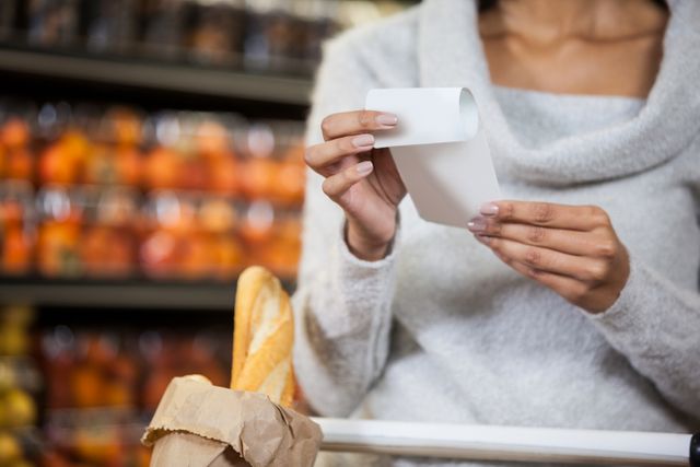 Woman holding and examining a receipt in a grocery store, with a shopping cart containing a baguette. Useful for themes related to shopping, budgeting, consumer behavior, retail, and grocery shopping tips.