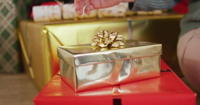 Golden gift wrapped box with a bow sitting on a red present in holiday setting with more gifts in the background. Ideal for holiday promotions, Christmas and New Year celebrations, festive greeting cards, and advertisements related to gift-giving.