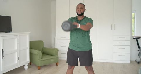 African american man performing kettlebell swings in home gym with smartwatch and earbuds