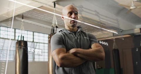 Bald muscular boxing trainer standing with arms crossed in industrial gym with hud overlay