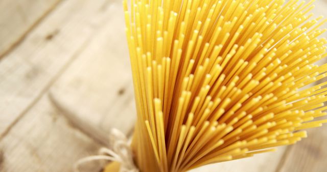 Close-up view of uncooked spaghetti stalks, tied together, standing on wooden table background. Suitable for use in food-related designs, Italian cuisine promotions, recipe books, or culinary blogs focusing on pasta dishes. Emphasizes the texture and color of the pasta against rustic wooden surface.