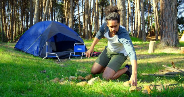 Young man arranging stones to create a campfire ring near his tent in a forest area. Scene captures the essence of camp setup, outdoor survival skills, and preparedness for an overnight stay in nature. Perfect for use in articles about camping tips, outdoor adventure, nature retreats, wilderness survival, and summer activities.