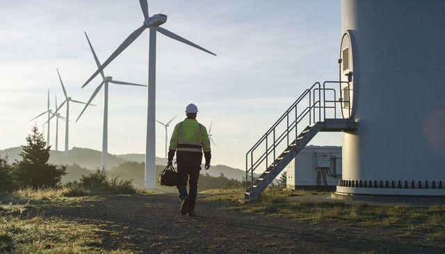 Wind farm technician walking toward turbine base at sunrise carrying tool bag and gear