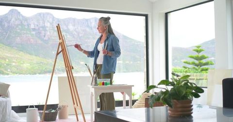 Mature woman painting on easel in bright lakefront studio with floor-to-ceiling windows