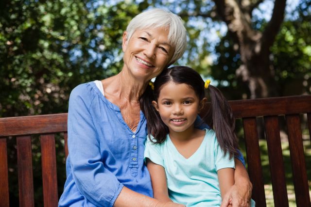 Portrait of smiling girl and grandmother sitting on wooden bench at backyard