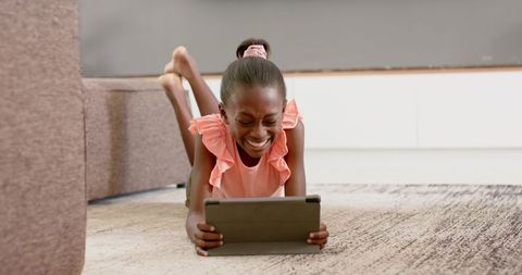 Smiling girl laughing while using tablet on living room rug, cozy family technology moment