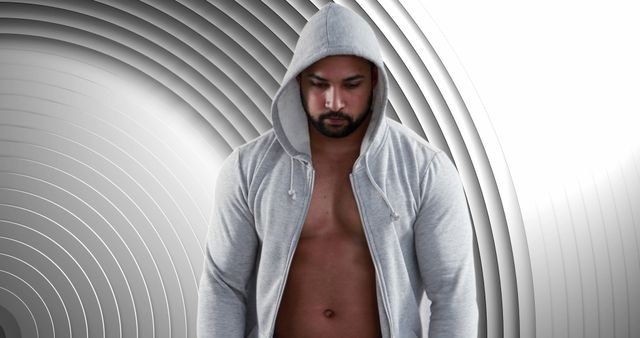 A confident man standing with his head slightly bowed and wearing a light grey hoodie. The modern geometric background features a pattern of grey arcs, giving the scene a futuristic and stylish feel. Perfect for use in fashion promotions, advertisements for men's activewear, or lifestyle blogs focused on contemporary men's fashion.