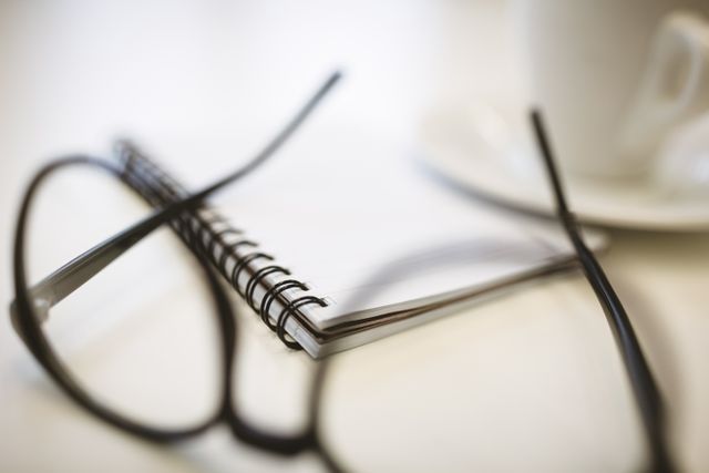 This image shows a close-up view of a notepad with eyeglasses resting on a desk in a creative office environment. A coffee cup is also visible in the background. This image is perfect for illustrating concepts related to productivity, planning, and work in a creative or professional setting. It can be used in articles, blog posts, or marketing materials focused on business, office life, or creative workspaces.