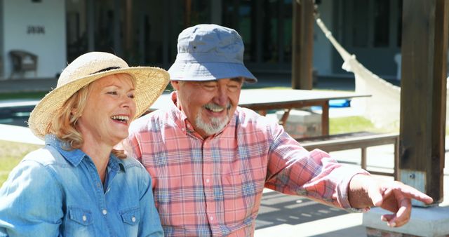 A senior Caucasian couple is sharing a joyful moment outdoors, with the woman wearing a straw hat and the man in a blue cap. Their laughter and pointing gesture suggest they are enjoying a lighthearted and engaging conversation.