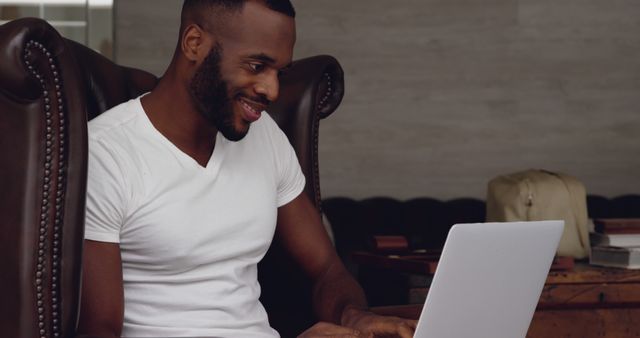 Man smiling and working on a laptop in a comfortable living room. Great for illustrating remote work, home office environments, productivity, and casual leisure at home.