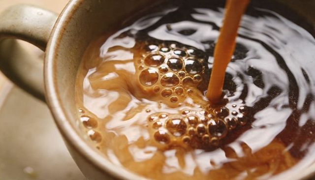 Pouring dark coffee into ceramic mug creating swirling foam bubbles and glossy reflection