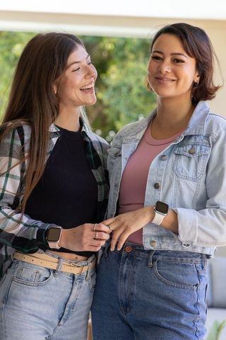 Diverse female friends laughing while touching fingers on sunny porch wearing smartwatches