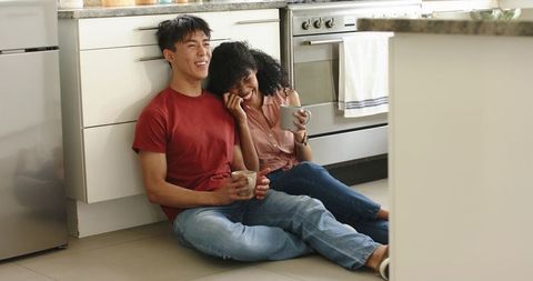 Asian man and african american woman sitting on kitchen floor, sipping coffee and laughing