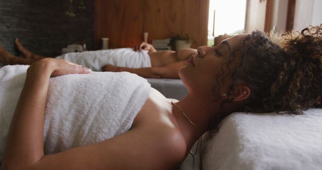 Image of relaxed diverse couple in towels lying with eyes closed on massage tables at health spa. Vacation, togetherness, relaxation, health, happiness and inclusivity concept.