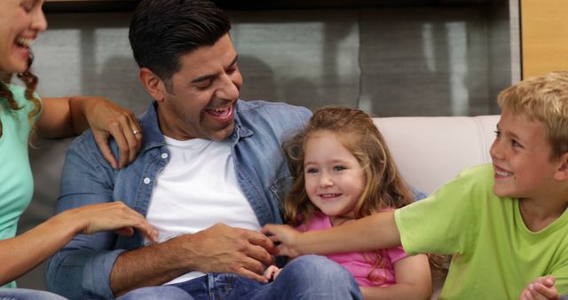 Happy family spending quality time together on couch, parents tickling children while everyone laughs. Picture captures joyful bonding and playful interaction, perfect for use in family-oriented marketing material, advertisements for family services, or parenting blogs illustrating the importance of family time.