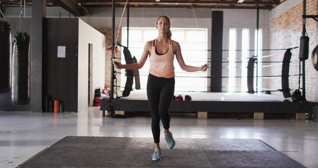 Young woman performing cardio exercise by jumping rope in a gym. A boxing ring is visible in the background. Ideal for articles on fitness routines, health tips, and gym equipment promotions.