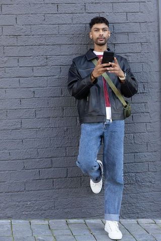 Man standing against gray brick wall holding smartphone wearing leather jacket and jeans