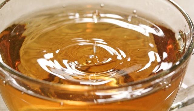 Macro closeup of amber tea droplet creating ripples in clear glass cup with warm reflections
