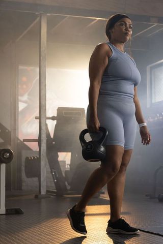 Backlit african american woman training with 5kg kettlebell in gym wearing blue active set