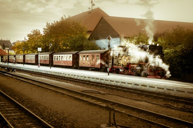 Shows a vintage steam train stopped at a station platform in a nostalgic setting. Historical and old-fashioned scene with added charm of steam and iron tracks, enhanced by autumn foliage. Ideal for travel promotions, historical documentaries, railway enthusiasts, or articles about nostalgic journeys and transportation history.