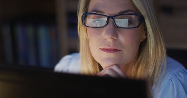 A woman with glasses concentrating intensely while working at a computer late at night. The reflection of the screen can be seen in her glasses. Ideal for illustrating concepts related to productivity, dedication to work, office environments, night shifts, hard work, remote work, and professional career development.
