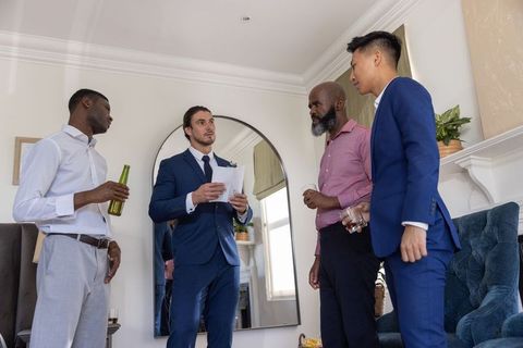 Diverse groomsmen gathering at home with navy-suit man reading speech and toasting