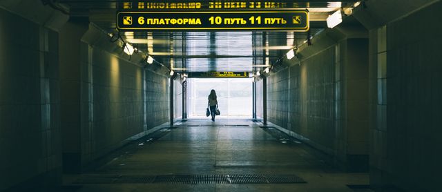 Image showing a woman walking through a tunnel in a train station. The tunnel is lit with overhead lights and displays directional signs in Russian. Suitable for depicting urban travel, transportation, and solo journeys.