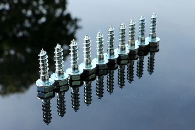 Metal screws lined up in a row reflecting on a shiny surface. Suitable for illustrations related to construction, hardware supplies, home improvement projects, engineering, and industrial work environments. Ideal for promotional materials for hardware stores or educational resources on tool usage.