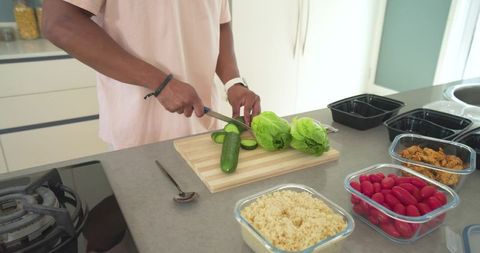 African american man chopping cucumber and packing meal-prep containers in modern kitchen