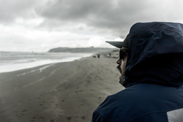 A man in a blue jacket and sunglasses is standing on a beach with overcast skies and the ocean in the background. He appears to be deep in thought while watching the waves roll in. Ideal for use in themes of contemplation, solitude, nature, travel, or moody weather conditions.