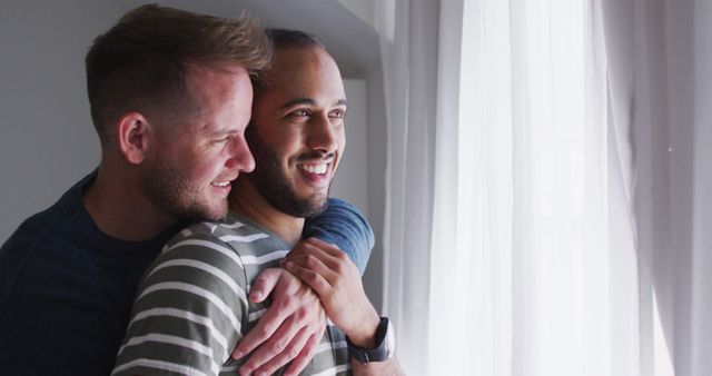 Two men in a loving embrace are enjoying a joyful moment near a window and bathing in natural light. Perfect for depicting concepts of love, LGBTQ relationships, and bonding in promotional materials, social media content, or lifestyle blogs.