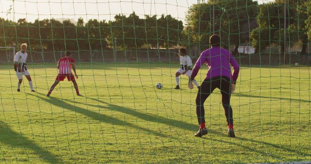 Soccer players engaged in a competitive match on a grassy field, viewed from the perspective of a goalkeeper. The game is taking place during sunset, creating dramatic lighting. This image can be used for sports promotions, articles about soccer, and fitness-related content.