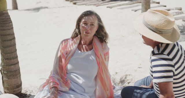 Senior couple sitting on sandy beach enjoying sunny day. Man wearing hat and striped shirt, woman in light dress with draped scarf. Discussing their day in serene vacation setting, ideal for promoting travel destinations, retirement living, and outdoor activities.