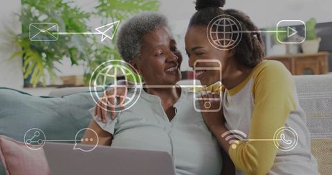 Adult daughter teaching mother to use laptop on cozy sofa with digital icons overlay