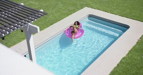 African american woman relaxing on pink inflatable ring floating in backyard pool