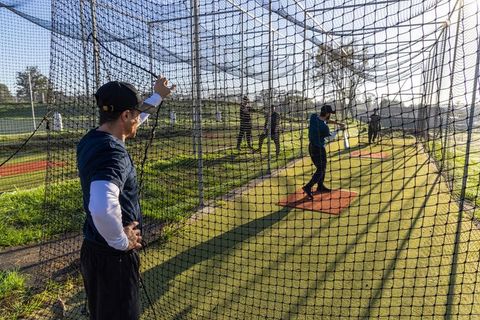 Coach guiding batter swinging during outdoor batting cage practice on turf at sunset