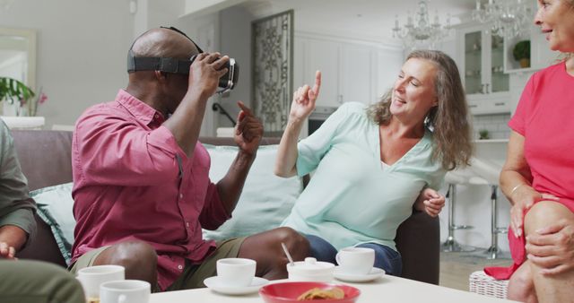 Diverse group of friends sitting in a cozy living room, enjoying virtual reality and having fun together. Seen holding VR goggles and laughing, engaging in a playful and interactive social activity. Perfect for advertisements or blog posts related to technology, senior lifestyle, digital connectivity, or group activities for older adults.