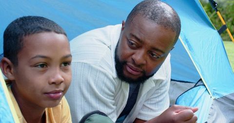African american father watching teen son camping in blue tent, bonding outdoors
