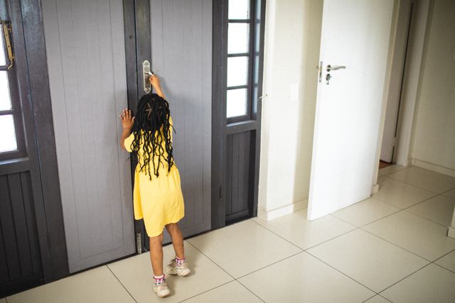 This image shows an African American girl with braided hair wearing a yellow dress, reaching up to open a door in a home. It captures a moment of childhood curiosity and exploration. This can be used in articles or advertisements related to parenting, child safety, home life, or childhood development.