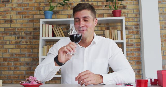 A man in a white shirt smiling while holding a glass of red wine and sitting at a table indoors. Suitable for concepts related to leisure, relaxation, leisure activities, indoor settings, and lifestyle promotions.