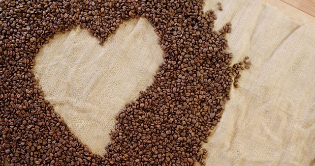 Coffee beans are arranged in a heart shape on a burlap surface, with copy space. It's a creative display that suggests a love for coffee or could be used for a romantic concept involving the popular beverage.