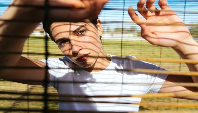 Young woman peering through bamboo blinds casting striped shadows across face in warm outdoor light