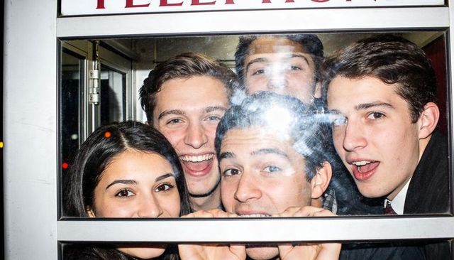 Laughing friends pressing faces to glass inside phone booth during lively urban nightlife