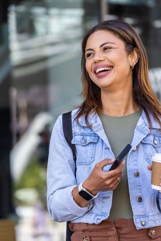 African american woman smiling while holding smartphone and coffee on urban sidewalk
