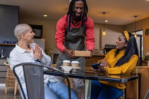 Smiling black barista handing takeaway box to delighted friends at cozy cafe table