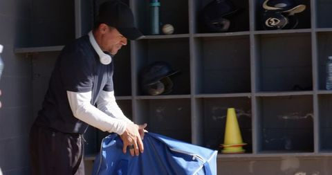 Baseball coach packing equipment bag in sunlit dugout with helmets and cones