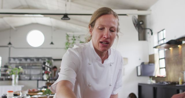 Female chef is cooking in a well-equipped modern kitchen wearing a white uniform. She appears focused and dedicated to her craft. This stock photo can be used for content related to cooking tutorials, restaurant advertisements, culinary schools promotions, or lifestyle blogs focused on gourmet cooking.