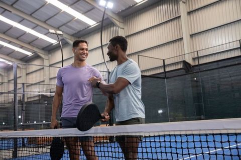 Smiling diverse male friends celebrating win on indoor pickleball court holding paddles